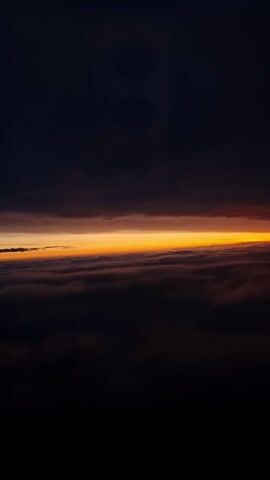 The video displays a tranquil view from above the clouds. A vibrant orange horizon separates the dense cloud layer below from the dark, upper sky. There are no visible people or objects in the video, the focus being solely on the atmospheric view. The environment conveys a sense of calmness, with the only action being the subtle movement of the clouds. There are no clear interactions within the frame, emphasizing the vastness and serenity of the scene.