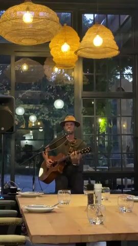 The video features a man playing an acoustic guitar and singing into a microphone in an indoor dining area. He is wearing a hat, a green T-shirt, and dark pants. There is a speaker to his left and a table in front of him set with plates, glasses, and condiments. Above, there are several rustic-looking woven light fixtures. The background includes large windows through which greenery and illuminated outdoor lighting can be seen.
