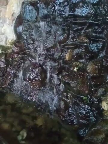 The video shows a close-up, top-down view of a small artificial waterfall or water feature. Water is flowing over rough, dark-colored stones and a tiered structure, creating a cascading effect. The water pools in a green-tinged basin below. The surrounding environment appears to be composed of rocks and possibly some vegetation, suggesting an outdoor setting. The lighting is dim, making it difficult to discern fine details. No people or animals are visible in the footage.