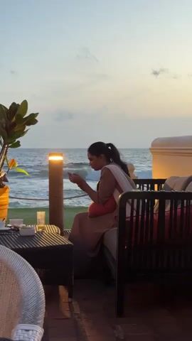 The video shows an outdoor restaurant setting with a seaside view at dusk. The sky transitions from light to dark blue, and the ocean is visible in the background. Several people are seated at tables, most of them are looking at their phones. There are menus on a wicker table in the foreground. The seating area is decorated with lights, plants, and cushions. The overall atmosphere appears relaxed and scenic.