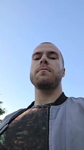 A man walks down a paved road lined with trees and greenery. He is wearing a gray jacket over a t-shirt and has a beard and a shaved head. A small black and white dog trots ahead of him on the road. The sky is blue and clear. The video is filmed from the man's perspective.