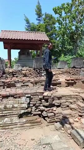 A person walks among ancient brick ruins under a clear blue sky. The ruins appear to be part of a temple or historical site, with crumbling walls and some large stone blocks. In the background, a covered structure shelters a large stone statue. Trees and foliage surround the area. The person, wearing a dark jacket and jeans, moves through the site, exploring the remnants of the past.