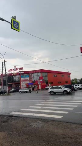 This is a medium shot video of a street scene. The camera is positioned looking towards a large, red building with the word APELSIN in large letters across the top. In front of the building is a pedestrian crossing, and several cars are visible. The sky is overcast and it appears to be raining, as the road surface is wet and reflective. A pedestrian crossing sign is visible hanging from a pole on the left side of the frame. A white SUV is driving across the pedestrian crossing.