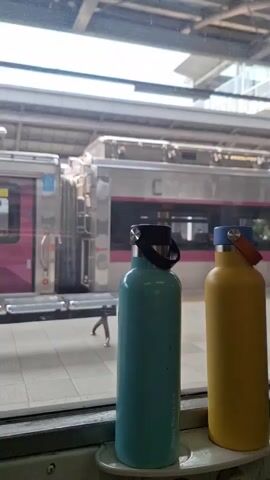 The video shows two water bottles, a teal one and a yellow one, sitting on a shelf in front of a window at a train station. The train station platform and a train with a pink stripe are visible through the window. The train is stationary. The teal bottle has a black handle and the yellow bottle has an orange handle. The bottles are labeled "Le Ding Ding" on the bottom. People can be seen walking on the train platform. The video is steady.
