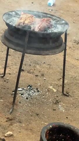 The video shows raw meat cooking on a makeshift grill. The grill appears to be made from an old tire rim supported by rebar legs. There are two pieces of meat on the grill, with smoke rising from them. The ground is dirt and scattered with debris, including a plastic water bottle and what looks like trash. In the background, two dogs are visible, one standing and the other bent over. The lighting suggests it is daytime.