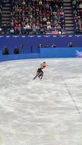 A figure skating duo is performing in an indoor arena with a large crowd watching. They are performing on a large ice rink surrounded by blue barriers displaying the Milano Cortina 2026 logo and the Olympic rings. The male skater is dressed in black while the female skater is dressed in black with gold accents. They are performing a synchronized routine with spins, glides, and coordinated movements. There are cameras and photographers present, documenting the performance. The crowd is densely packed in the stadium seating.