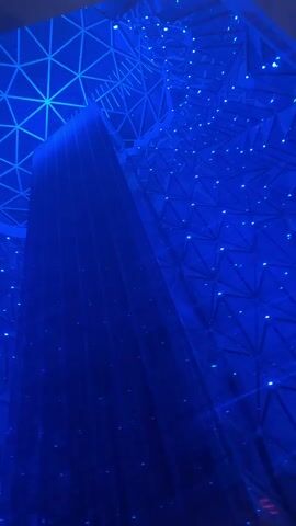A geodesic dome ceiling, illuminated by changing colors of light, is visible. The dome is composed of triangular facets, and small points of light are scattered across its surface. In the center of the dome, a tall, dark, rectangular structure extends upwards. The lighting transitions through shades of blue, purple, red, and green.