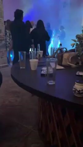 The video shows a scene at a party. People are dancing in the background, illuminated by blue and green lights and haze. In the foreground is a dark, round table with several empty glasses, a white cup, and a small decorative object. There's also a handbag on the table. The room has a stone wall visible on the left, and the floor appears to be tiled. The focus is on the table and its contents, with the dancing figures slightly blurred in the distance.