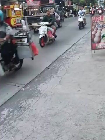 A street scene captured from a low angle looking up towards a road with traffic. The foreground is a cracked concrete surface. On the road, several motorbikes are visible. One motorbike, white with red accents, is in the left foreground with two people riding it, the driver wearing a white hat and the passenger with a white backpack. Further down the road, other motorbikes and a pickup truck are traveling. On the left side of the road, there are food stalls, one with a yellow and red facade. On the right side of the road, a sign with pictures of eggs is visible. The sky is overcast. The overall impression is a busy street in a populated area.