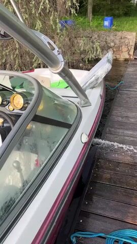 The camera frames a white and maroon boat docked on a wooden pier. The boat has a steering wheel and several gauges visible through the windshield. A silver metal frame with mounted clamps is positioned above the boat, and a section of the boat is covered in clear plastic. Water splashes between the boat and the pier. In the background, there are trees, bushes, and blue containers.