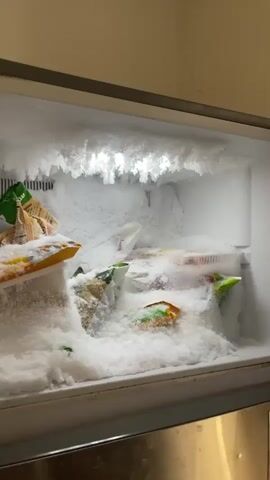 The video displays the interior of a heavily frosted freezer. Thick layers of white ice and crystalline frost cover the ceiling, walls, and floor of the appliance. Several plastic bags, possibly containing frozen food, are partially buried within the accumulation of ice. Large, jagged ice crystals are particularly prominent on the freezer's ceiling, illuminated from within. The video starts with a wider view of the frozen interior and then zooms in to showcase the intricate ice formations. Conversational speech in an unidentified language is audible throughout the recording.