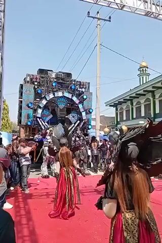 The video shows a procession or parade with performers in elaborate costumes. In the foreground, two individuals are wearing large, dark, winged headdresses and long, ornate dresses. The person on the right has long, reddish-brown hair. They are walking on a red carpet laid out on a paved surface. Behind them, a large, multi-tiered sound system or stage setup is visible, decorated with lights and intricate designs. Several people are gathered around the sound system, some holding cameras or phones. Further back, on the right side of the frame, a building with green and white details, including a dome, is visible, resembling a mosque. The sky is clear and bright, indicating daylight. The overall scene suggests an outdoor event, possibly a festival or celebration, with a musical or performance element.