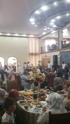 The video shows an indoor gathering, likely a celebration, with people seated at tables laden with food. In the background, a group of men dressed in traditional attire, including hats, stand together. One man holds a long, brass instrument. Other individuals are seen entering or exiting through an arched doorway. The environment appears to be a banquet hall with ornate curtains and a decorative ceiling.