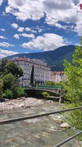 The video shows a river flowing in the foreground with rocks and clear water. A bridge crosses the river, with a large, ornate building behind it. The building is light-colored with many windows and a red-tiled roof. Mountains are visible in the background, and the sky is blue with fluffy white clouds. Trees line the riverbanks. The overall scene appears to be a scenic landscape in a town or city. No people or specific actions are visible.