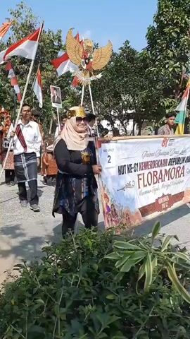 A group of people are participating in a parade. Several individuals are holding flags, including the Indonesian flag, and some are carrying banners. One banner reads "HUT KE 61 MERDEKA FLORES". The people are dressed in a variety of clothing, some in traditional attire and others in more casual wear. Some individuals are wearing face coverings or sunglasses. The parade appears to be taking place outdoors on a sunny day, with trees and foliage visible in the background. The participants are walking along a paved pathway. One person is carrying a framed portrait of a man. Another person is holding a large, stylized representation of a bird. Some participants are wearing elaborate costumes.