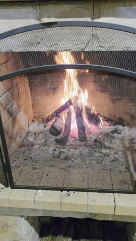 The video shows a fireplace with a fire burning inside. The fire is behind a black metal mesh screen. There are several logs in the fire, and flames are visible. There is ash and other debris on the floor of the fireplace. The fireplace is made of brick and stone. Sounds of people talking can be heard.