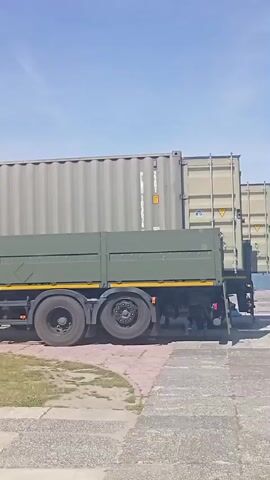 A green military truck is moving to the right. The truck is carrying a large, light-colored shipping container. The truck is on a paved surface with grass to the left. The sky is blue with some white clouds. In the background, there is a body of water with a few ships and a white car on the road.