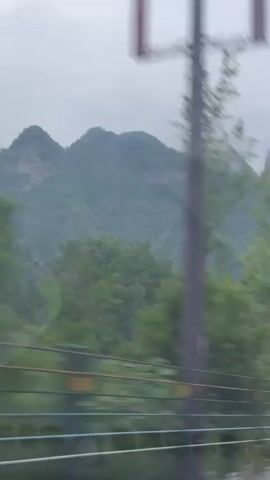 The video shows a view from inside a car traveling on a road with a mountain range in the background. The mountains are shrouded in mist and the sky is overcast. Green trees line the side of the road and a guardrail separates the road from the foliage. Street lamps are visible along the road.  As the car moves forward, a white car and a blue concrete mixer truck come into view, driving in the same direction. The video ends with the truck centered in the frame.