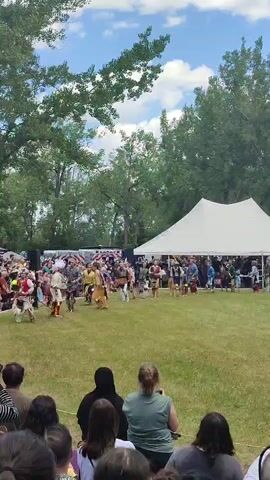 The video shows an outdoor event with a large crowd gathered on a grassy field. Several individuals are walking in a procession, some in traditional Native American regalia, including feathered headdresses and colorful clothing. They are walking on the grass in front of a large white tent and a line of trees. The sky is blue with some white clouds. The crowd in the foreground is watching the procession. The overall scene appears to be a cultural festival or powwow.