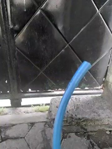 A close-up shot captures a blue hose moving horizontally. The hose is in focus and appears to be in motion, swinging from left to right. Behind the hose, a dark, possibly metal gate or fence is visible, featuring a diamond-patterned design. The lower portion of the scene reveals a stone or concrete surface, likely a walkway or ground, with some debris and patches of green grass or weeds peeking through the cracks. The overall lighting suggests an outdoor environment during daylight hours.