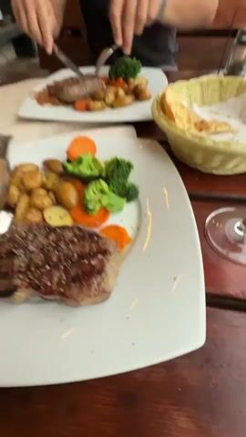 The video begins with a close-up shot of two plates of food on a wooden table. One plate contains a steak with potatoes, broccoli, and carrots. The other plate, slightly out of focus, also appears to have a steak with similar accompaniments. A basket of bread is visible in the background, along with a glass of red wine. The scene seems to be an outdoor dining setting, with a blurred background of lights. The perspective then shifts to a young woman with long brown hair, wearing a checkered jacket, who is filming herself. She is seated at a table, and in the background, a cat is visible near another table with food. The woman looks towards the camera and then to her side. The video continues with shots of the woman and her surroundings, including other diners and the restaurant environment. The setting appears to be a restaurant with outdoor seating, characterized by wooden furniture and overhead lighting. The woman interacts with the camera and her environment, creating a personal vlog-style video.