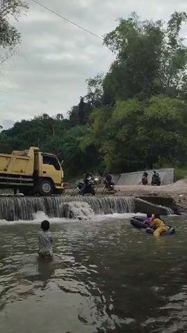 A dump truck is positioned on a concrete dam where a waterfall flows into a river. A person stands in the shallow water in front of the waterfall. To the right, two people are in the river, one on an inflatable tube. Several motorcycles and people are gathered near the dam. Lush green trees and foliage form the background under a cloudy sky.