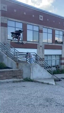 A group of people are gathered outdoors in a parking lot. In the foreground, a person on a scooter jumps down a set of stairs. Another person on the ground is filming. In the background, there are buildings and trees. Another person on a scooter appears to be performing a trick and falls. A person with a megaphone approaches the fallen individual.