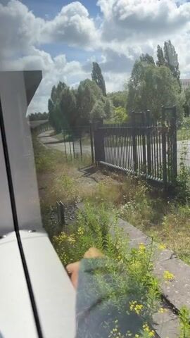 The video shows a person's hand gripping the edge of a white seat inside a moving vehicle, possibly a train. The camera is positioned to look out the window. Through the window, a fence, trees, and a pathway are visible. The vehicle moves past a green barrier with a striped pattern. The reflection in the window shows the back of a person with gray hair wearing a purple jacket, seated in front of the camera. The hand gripping the seat appears to be wearing a gray jacket or sleeve. The lighting suggests it is daytime.