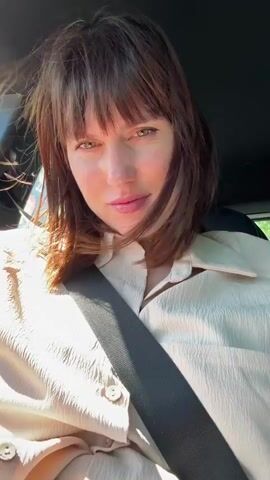 A person with light skin and brown hair with bangs is seated in the driver's seat of a car. They are wearing a light-colored, button-down shirt and a seatbelt. The person smiles slightly, and their hair is a bit tousled. They raise a hand to their hair, brushing it back and to the side. The car appears to be in motion, with sunlight and foliage visible through the windows. The car's interior features black leather seats and trim.