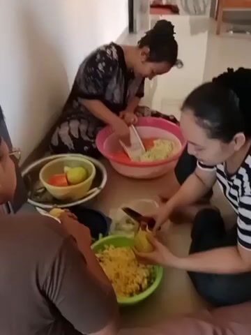 Three people are sitting on the floor, preparing food. Two women, one in a patterned dress and the other in a striped shirt, are in the foreground. The woman in the patterned dress is using a grater in a pink bowl filled with shredded vegetables. The woman in the striped shirt is holding a knife and peeling a yellow fruit over a green bowl of chopped ingredients. A third person, partially visible in the lower left, is also holding a yellow fruit and appears to be peeling it or preparing it for cutting. There are several bowls and a plastic bag containing cut vegetables on the floor around them. The environment appears to be indoors, possibly a home, with a light-colored wall and floor.