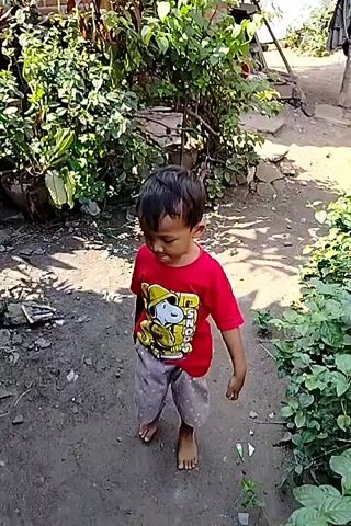 A young boy wearing a red t-shirt with a Snoopy graphic and gray shorts walks barefoot on a dirt path. Lush green foliage surrounds the path, with some rocks and debris visible. The boy is looking down as he walks.