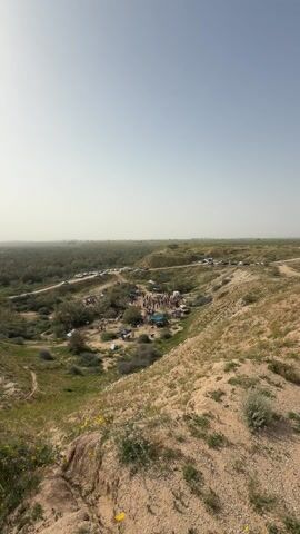 The video pans from a wide view of a valley to people standing on a hill overlooking it. 

The valley has a dirt road running alongside a treed area. Many cars are parked along the dirt road. 

In the center of the valley is an area with many people, tables, and possibly tents. 

The land is dry and grassy, with scattered yellow wildflowers. 

Three people stand on the hill at the edge of the video, facing towards the valley, looking down at the scene. One of the people is holding a red cloth.