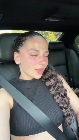 A young woman is in the passenger seat of a car, looking at the camera and speaking. Her face is flushed. In the background, a man is driving the car. The scene appears to be during the daytime, with trees visible through the car windows. The camera is filming from inside the car, moving slightly.