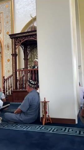A man in a white robe and cap stands at a wooden podium in a mosque, speaking to a group of people. A man in a grey shirt and kufi sits on the floor leaning against a white column. Another man in a white shirt and kufi sits behind him. Other men are also seated on the blue carpeted floor. The mosque has ornate gold calligraphy on the walls and a crescent moon symbol above the podium. There is a wooden stand with prayer beads on it.