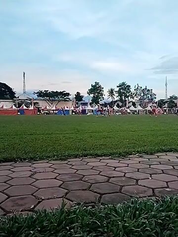 The video shows an outdoor scene, likely a public park or sports ground, in Matesih, Indonesia. In the foreground, there's a paved hexagonal path and lush green grass. The main area is a large green soccer field where a game is underway. Several people, dressed in soccer kits, are actively playing. One player in a dark jersey is seen attempting to kick the ball towards a white soccer goal, with a goalkeeper in a lighter jersey diving to defend.

In the background, along the far side of the field, multiple tents with white and red/orange striped canopies are set up, suggesting an event or market. Beyond the tents, there are numerous tall trees and some buildings, including one with a distinctive tall antenna or tower. The sky is a pale blue with some white clouds, indicating it's likely late afternoon or early evening. The overall atmosphere appears casual and community-focused.