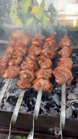 The video shows a close-up view of meat being grilled over hot coals. Several skewers of meat, some appearing to be chunks of beef or lamb and others chicken wings, are placed on a barbecue grill. Smoke rises from the grill, indicating the meat is cooking. The background is slightly blurred, showing greenery and possibly some outdoor seating. The lighting suggests it is daytime. The camera pans slightly across the skewers, offering a detailed view of the browning meat.