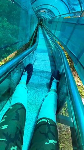 The video captures a first-person perspective of a person riding down a long, enclosed blue transparent slide outdoors. The primary subject, a female wearing sunglasses, a white t-shirt with an unreadable red graphic, and blue pants, records herself as she descends. Her feet, clad in blue slip-on shoes over white socks, are visible at the beginning, resting in the slide channel.

As the ride progresses, she turns the camera to show her face, revealing an expression of excitement and joy, with her mouth open in a shout or laugh. Another person, also female and wearing a white top, is visible riding the slide behind her. The slide enclosure, made of a translucent blue material, offers views of lush green vegetation and trees outside, suggesting a natural, possibly mountainous or hilly, setting in Jiejinkou, China. The activity takes place during the daytime, under bright and clear weather conditions. No discernible text is clearly visible other than the partial graphic on the primary subject's shirt.