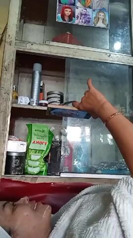 The video shows a person receiving a facial treatment in what appears to be a salon. A person's face is visible in the foreground, lying down and being attended to. Their face is covered with a white towel. In the background, a shelf unit is visible, filled with various beauty products and items. These include hairspray cans, jars, and what looks like a package of aloe vera gel. Above the shelf unit, there is a display of photographs featuring different hairstyles and hair colors. A hand with a bracelet is visible in the upper right portion of the frame, holding a smartphone.