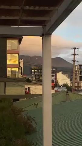 The video shows a rooftop view of a cityscape, framed by the metal structure of a balcony or shelter. The roofs in the foreground are mostly green and corrugated. Buildings of varying heights and architectural styles are visible, including some under construction. In the background, there are mountains partially obscured by clouds. The sky is a mix of blues and greys, with some pink and yellow hues from the setting sun. The camera slowly pans across the scene. There are utility poles with wires running through the shot.