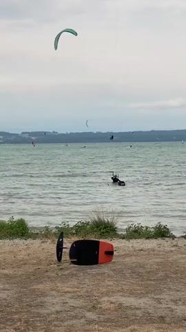 The video shows a body of water with several kitesurfers on it. In the foreground, a foilboard lies on the beach. The board is black and orange. In the background, several kitesurfers are visible on the water, with their kites in the air. One kitesurfer is in the water, appearing to be adjusting their gear. The sky is overcast.