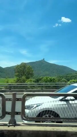 This video captures a scenic view from a moving vehicle, likely a car or bus, in Hoa Xuan, Vietnam. The primary focus is a lush green mountain with a distinctive, jagged rock formation at its peak, resembling a sculpted figure or a cap. The mountain is covered in dense vegetation, suggesting a tropical or subtropical environment.

In the foreground, we see a highway with white lane markings and a diamond-patterned shadow cast on the asphalt. A metal barrier runs along the side of the road, and beyond that, the foliage of trees. A white truck with the license plate "29K-052.31" is visible in the mid-ground, passing by. Another vehicle, possibly a car, is partially visible behind the truck.

The sky is a vibrant blue with a few wispy white clouds, indicating fair weather and likely daytime, possibly late morning or early afternoon given the brightness. The overall atmosphere is serene and picturesque, highlighting the natural beauty of the Vietnamese landscape. The camera's movement suggests a journey, offering glimpses of both the road and the distant mountainous terrain.