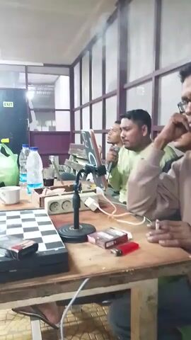 The video shows a group of men in a room. In the foreground, a chessboard is set up on a wooden table. There are also cigarettes, lighters, a microphone on a stand, a tablet, a cup, and water bottles on the table. One man is holding a cigarette and appears to be talking on the phone. Another man is holding a microphone and looking at the tablet. In the background, more men are seated around another table. There are also chairs, a green bag, and an exit sign visible. The room appears to be an indoor space with fluorescent lighting.