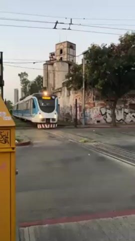 The video shows a train crossing a street. The train is painted in white, light blue, and black, and has red and white stripes on the front. The train is traveling from left to right. The train horn is heard. A yellow traffic barrier is in the foreground on the left. Behind the train is a building with graffiti on it. To the left of that building is a silo-like structure. In the background are trees and a tall building. The sky is a pale blue.