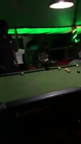 The video shows a dimly lit pool hall. Around a green pool table, several people are gathered. Some are seated on cushioned benches along the sides, while one person is actively playing pool, aiming and shooting the cue ball with a pool stick. The overhead lighting above the pool table alternates between red, green, and blue. The atmosphere appears casual and social.