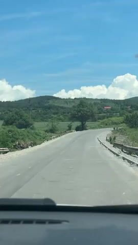 The video presents a bright, sunny daytime scene captured from inside a vehicle, looking out through the windshield onto a rural landscape in Baylovo, Bulgaria. The top of the car's dashboard is visible at the bottom of the frame.

A paved road stretches ahead, showing some signs of wear, and curves gently to the right. Along the right side of the road, freshly laid gravel and curb stones indicate recent construction or ongoing road work. Both sides of the road are lined with abundant green vegetation, including various bushes and trees. A large, prominent deciduous tree stands near the bend in the road on the left side.

In the midground and background, rolling green hills rise, densely covered with trees and fields. A few distant houses with red roofs can be spotted on the hillsides. Utility poles are also visible against the sky. The sky above is a clear, vibrant blue, adorned with scattered, fluffy white cumulus clouds. The strong sunlight suggests it is likely midday or early afternoon. No people or animals are visible in the scene.