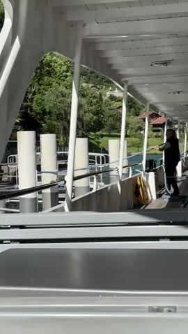 The video shows a view from a ferry. There are two individuals on the deck of the ferry, one of whom is holding a hose and appears to be using it. The ferry is docked at a pier, with white cylindrical bollards visible. In the background, there is a body of water, lush green trees, and houses on a hillside. The lighting suggests it is daytime.