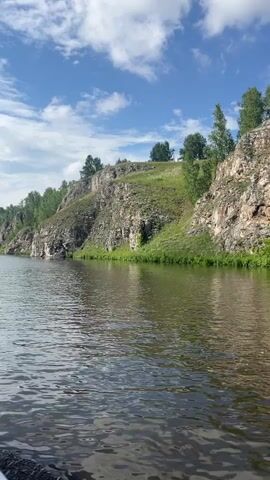 A first-person perspective video shows a boat moving on a river towards a rocky cliff face. The cliff is steep and covered in green vegetation at the top. Trees grow along the ridgeline. The sky is blue with scattered white clouds. The water is rippling and reflects the sky and cliffs. The front of the boat is visible in the lower left corner of the frame. The camera pans slowly to the right, following the curve of the river and the cliff.