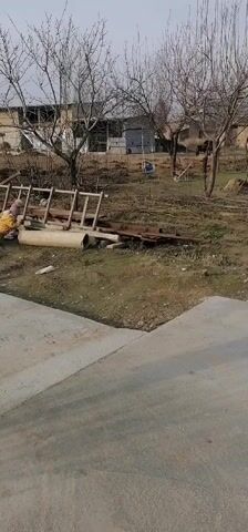 The video shows a rural setting on a day with overcast skies. In the foreground, there's a section of concrete pavement. Beyond the pavement, the ground is covered with grass, dried leaves, and miscellaneous debris.  

A child, wearing a yellow outfit, is seen playing near a pile of objects, including several large concrete pipes and wooden ladders.  

In the background are bare trees and simple buildings, possibly houses or barns. A swing hangs from one of the trees.  

The camera seems to be stationary, capturing the scene from a distance.