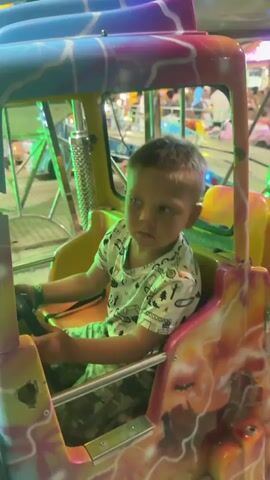 A young boy sits in the driver's seat of a colorful toy truck. The truck is part of an amusement park ride, with other rides and people visible in the background. The boy is wearing a t-shirt and camouflage shorts. He holds the steering wheel and looks around, his expression changing from a blank stare to a slight frown. The truck has a bright, psychedelic paint job with swirls of pink, purple, blue, and orange, resembling lightning bolts or flames. The ride appears to be in motion or preparing to move, as indicated by the background activity.