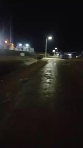 The video shows a dark, outdoor scene at night. A single streetlamp illuminates a wet, possibly muddy, path or road. The ground reflects the light. In the background, there are other lights that appear blurred, suggesting they are distant or moving. The overall impression is of a deserted area, possibly rural or industrial, under artificial lighting during nighttime hours. There are no people visible in the frame.